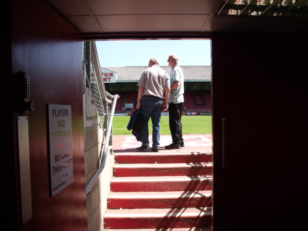 Zwei ältere Männer stehen vor einer Treppe in einem Stadion. 